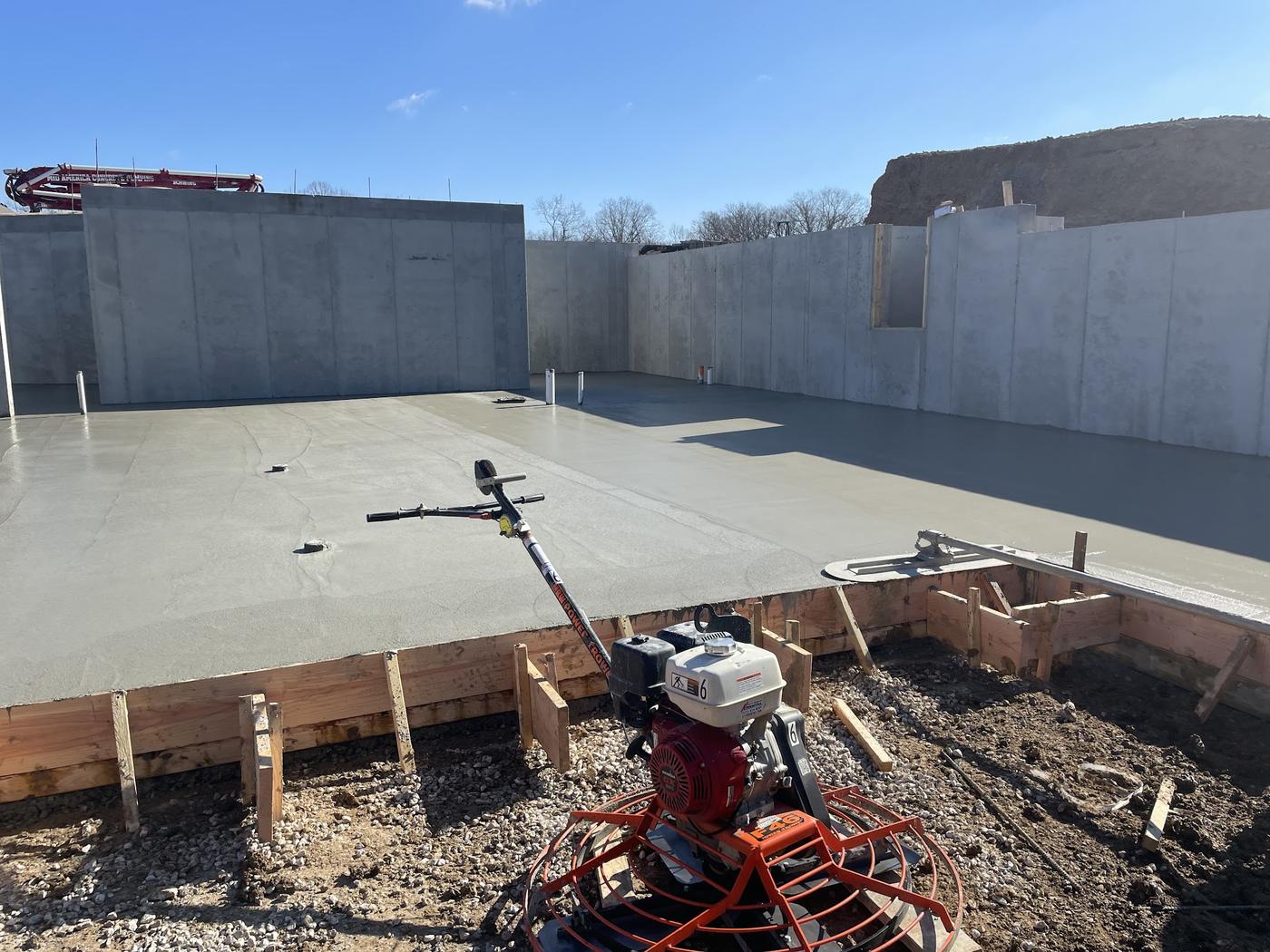 A finished basement slab under blue sky inside poured foundation walls, with a walk-behind power trowel resting at the edge